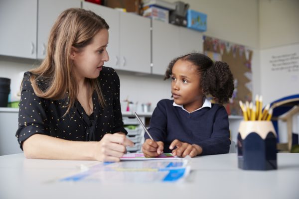 Young female primary school teacher working one on one with a schoolgirl at a table in a classroom, looking at each other, close up